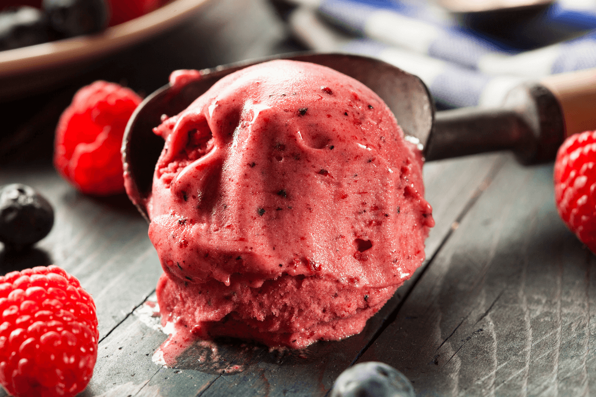 Eine Portion Beereneis in einer Schaufel, umgeben von frischen Himbeeren und Heidelbeeren auf einem Holztisch.