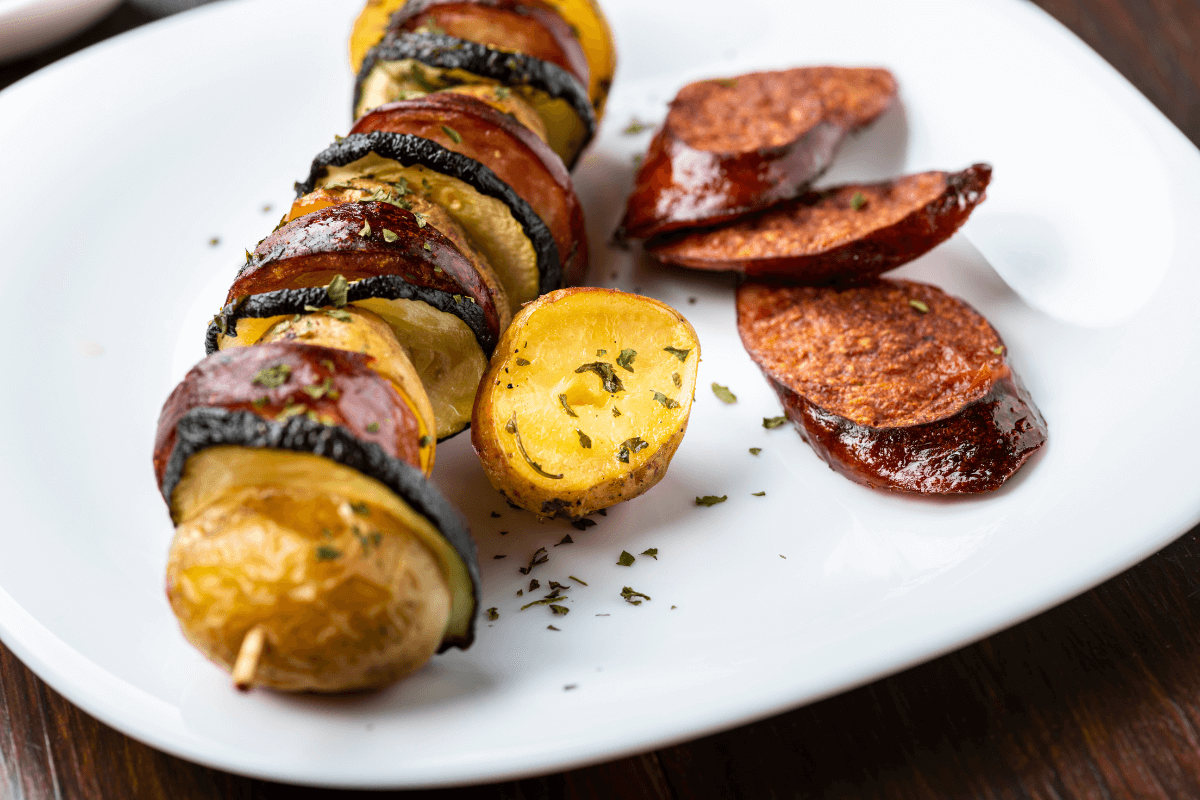 Gegrillte Kartoffel-Würstchen-Spieße mit Zucchini und knusprigen Wurstscheiben, garniert mit Kräutern auf einem weißen Teller.