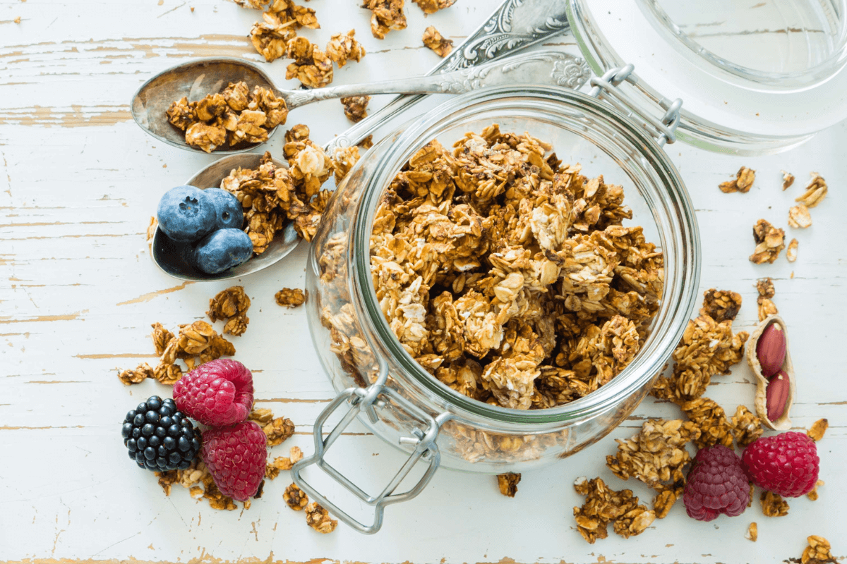 Glasgefäß mit knusprigem Granola, umgeben von frischen Beeren (Himbeeren, Heidelbeeren, Brombeeren) und Löffeln.