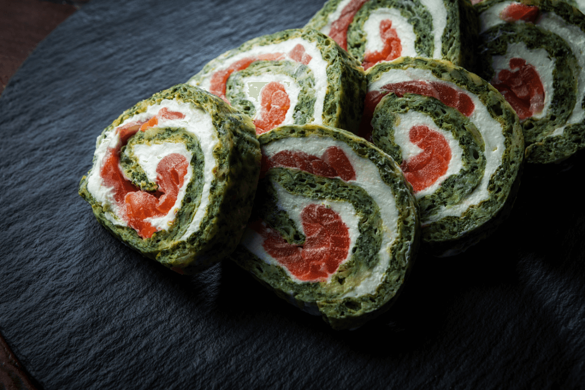 Frische Lachs-Spinat-Rolle, spiralförmig geschnitten, gefüllt mit Frischkäse und Lachs, auf einem schwarzen Servierbrett.