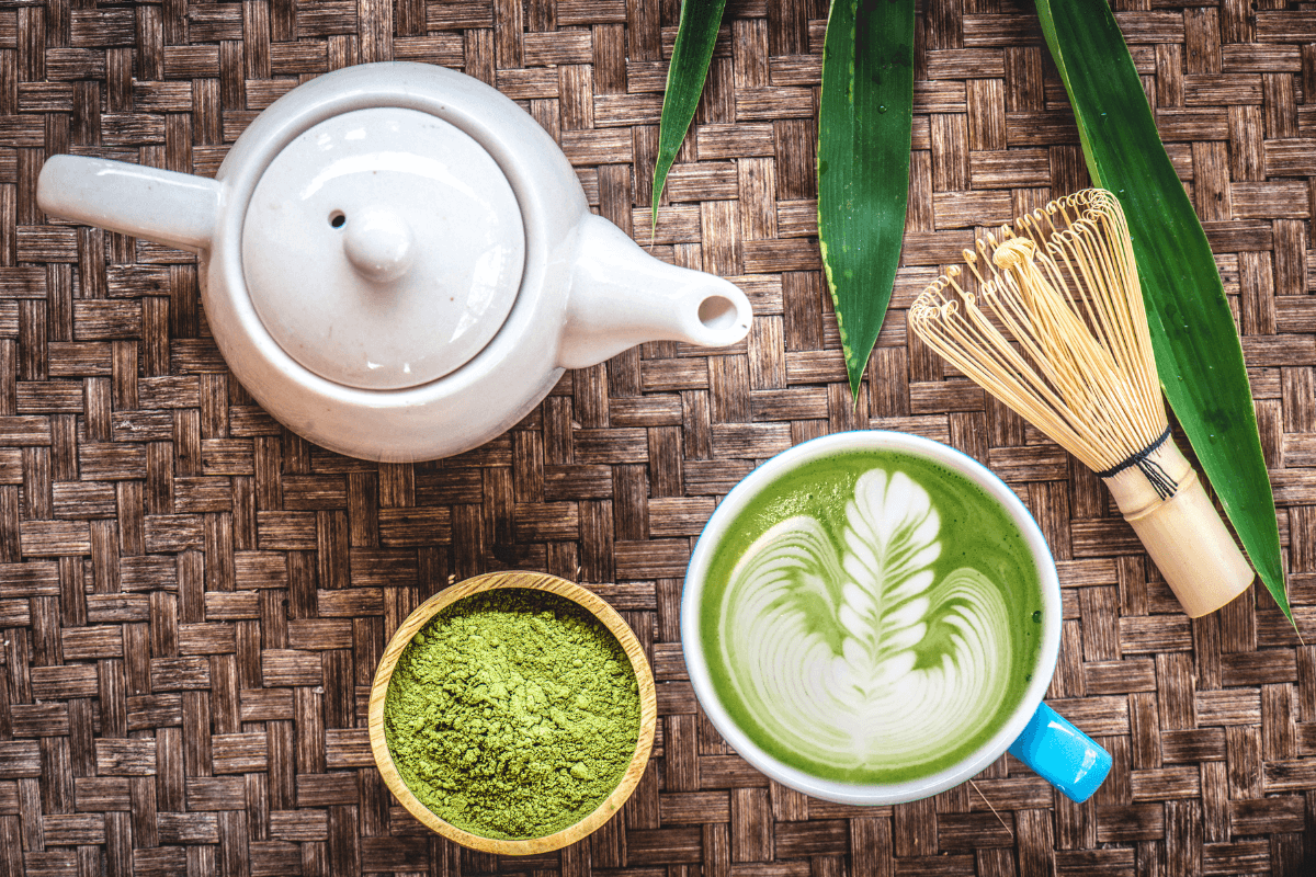 Teekanne, Matcha-Pulver, Matcha-Latte mit kunstvollem Muster und Bambusbesen auf einem geflochtenen Untergrund.