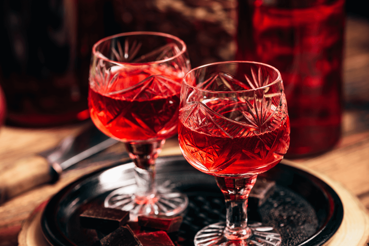Zwei dekorative Gläser mit rotem Rhabarberlikör auf einem schwarzen Tablett, umgeben von Schokoladenstücken.