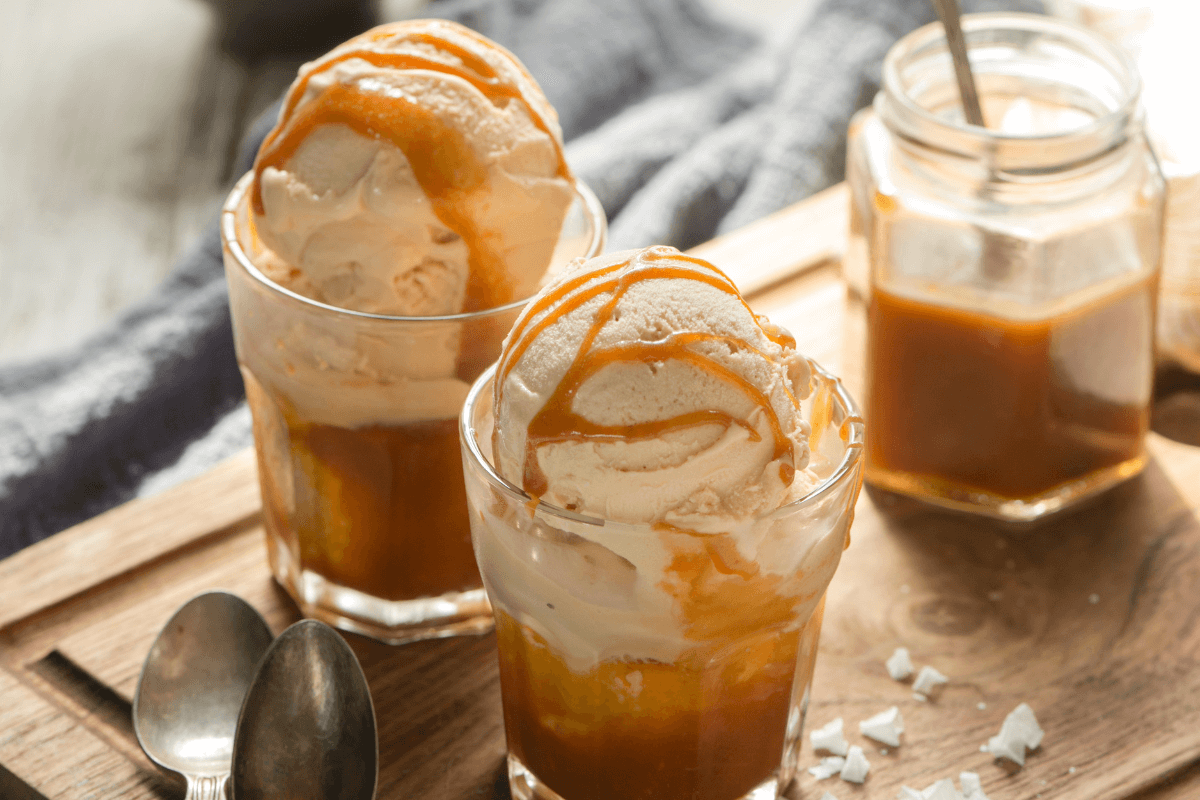 Zwei Gläser mit Vanilleeis, überzogen mit Karamellsauce, auf einem Holzbrett. Daneben ein Glas mit Karamelsauce.