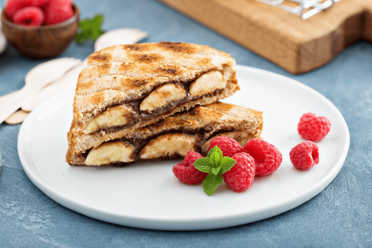 Schoko-Bananen-Sandwich auf einem weißen Teller, geschnitten, mit frischen Himbeeren und Minze dekoriert.