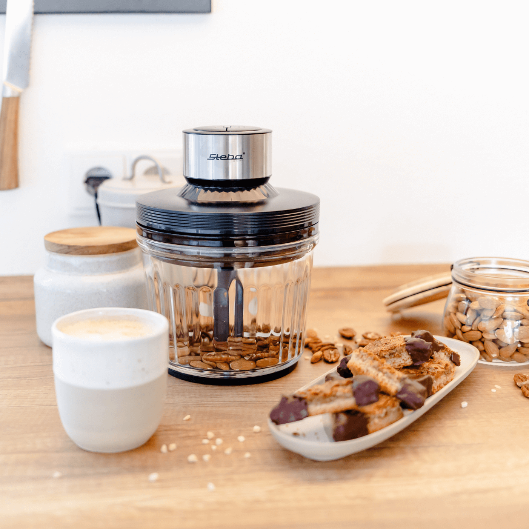 Steba Elektrogerät zum Mahlen von Nüssen und Gewürzen, umgeben von Kaffeetasse, Keksen und einer Glasdose mit Mandeln.