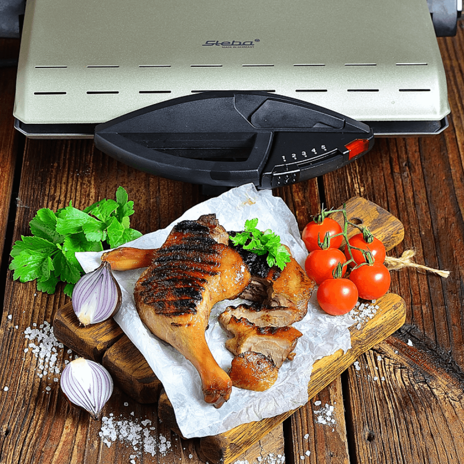 Kontaktgrill von Steba mit gegrilltem Hähnchenschenkel, frischen Kräutern, Zwiebeln und Tomaten auf einem Holzbrett.