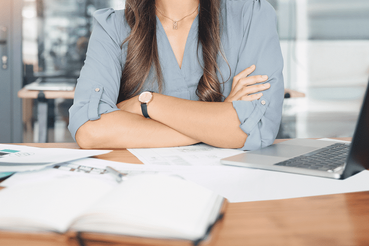 Eine Person mit verschränkten Armen sitzt an einem Schreibtisch mit Laptop und Dokumenten, in einer modernen Büroumgebung.