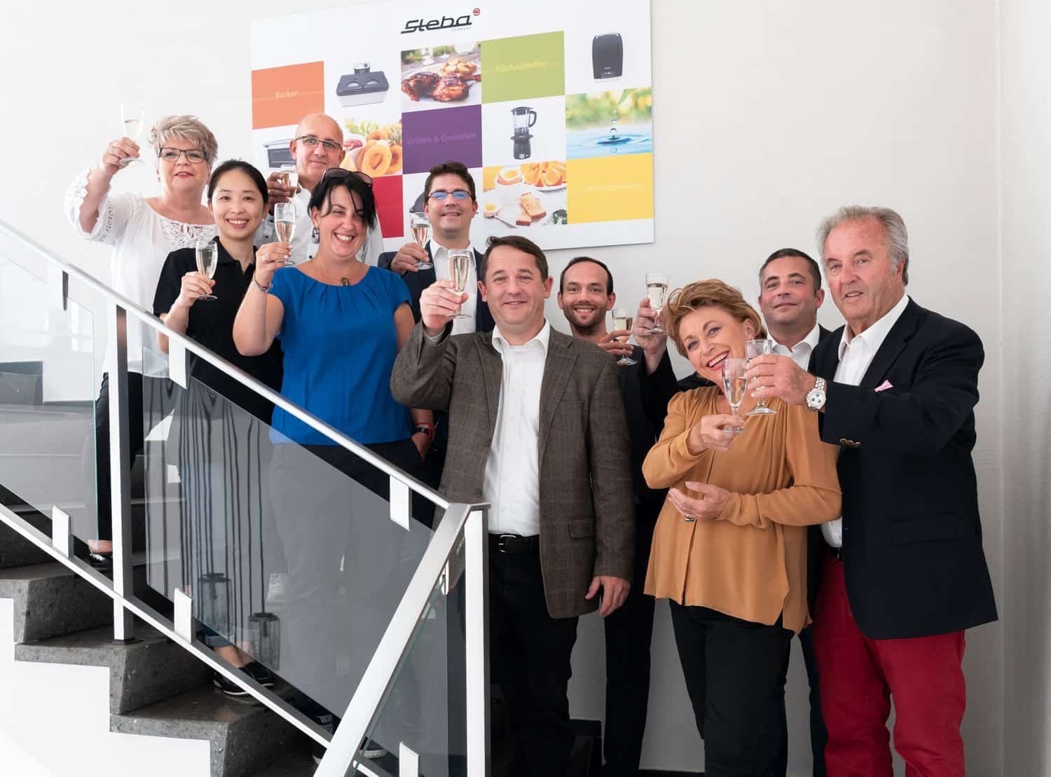 Gruppierung von Personen, die auf einer Treppe mit Sektgläsern anstoßen, im Hintergrund Steba-Wand mit Elektrogeräten.