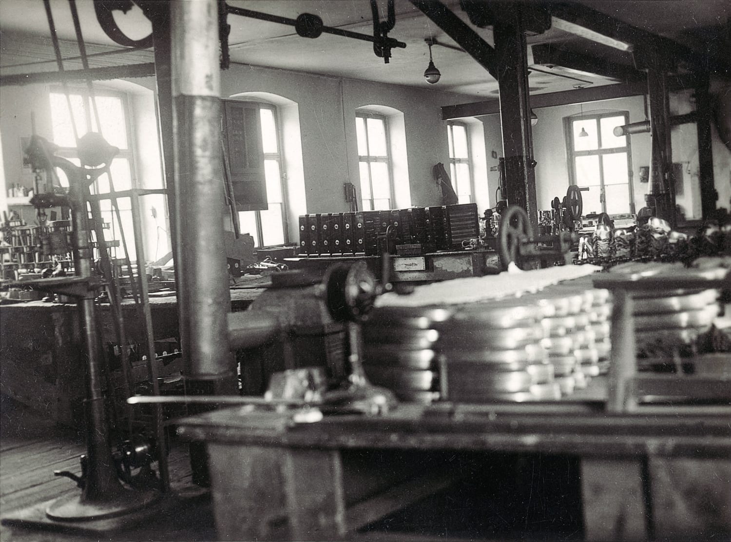 Historische Montagehalle mit Maschinen, Werkzeugen und gestapelten Metallteilen. Helle Fenster sorgen für natürliche Beleuchtung.