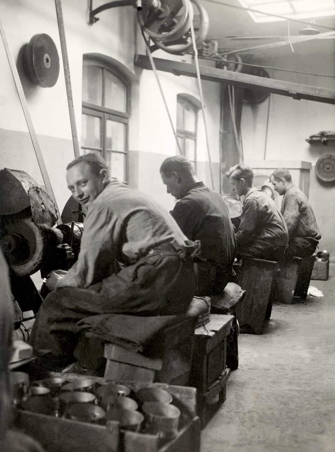 Arbeiter in einer Poliererei um 1900, sitzen an Werkbänken und bearbeiten Metallteile mit Maschinen.