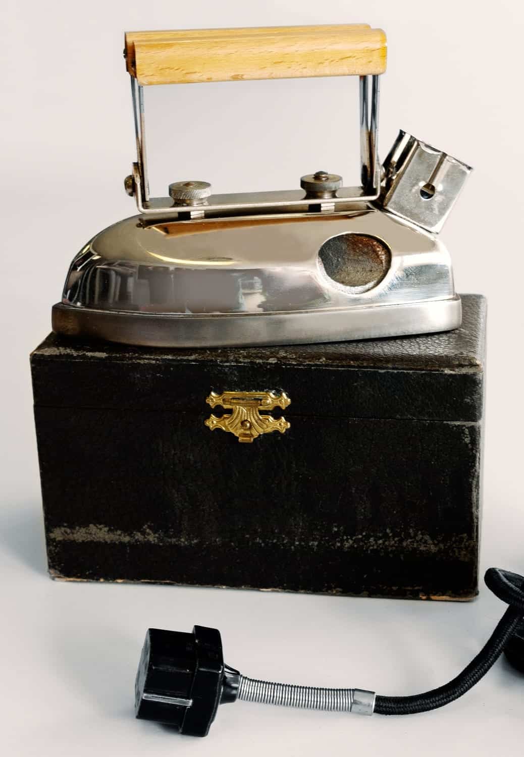 Reisebügeleisen mit Edelstahl-Oberfläche und Holzgriff, auf schwarzem Koffer mit goldener Schnalle. Kabel sichtbar.