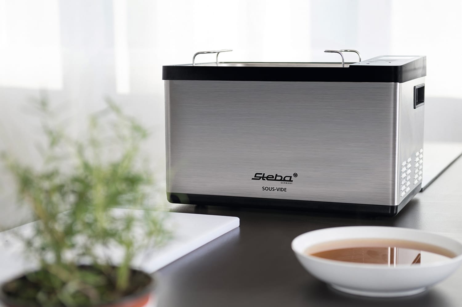 Sous-Vide Gerät von Steba in Edelstahloptik mit schwarzem Deckel, auf einem Tisch neben einer Schale mit Flüssigkeit.