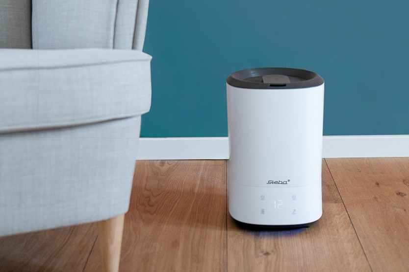 Luftbefeuchter Steba LB 8 in modernem Design, platziert auf Holzboden neben einem grauen Sessel, mit digitaler Anzeige.