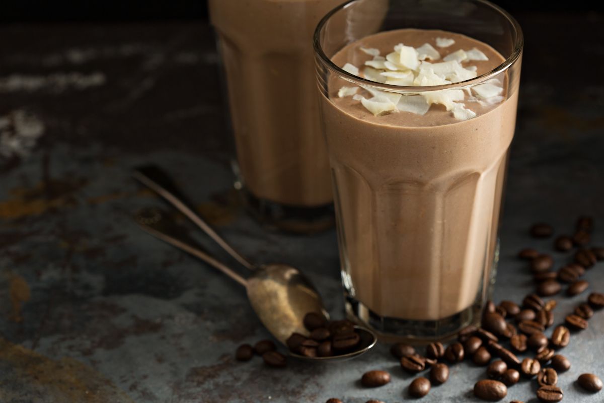 Zwei Gläser Kaffee-Bananen-Smoothie, garniert mit Kokosraspeln, neben Kaffeebohnen und Löffeln auf einer dunklen Oberfläche.