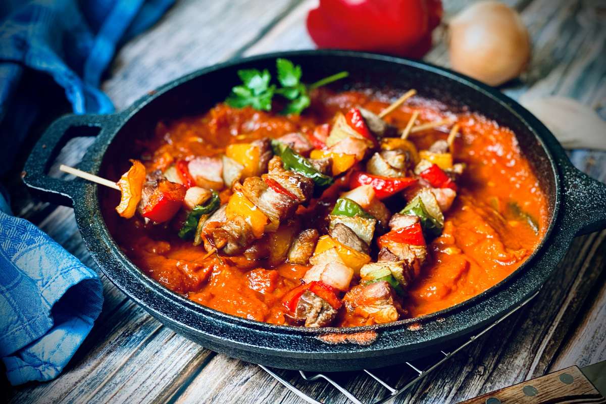 Frittierte Schaschlik-Spieße in einer schwarzen Pfanne mit Tomatensauce, umgeben von buntem Gemüse.