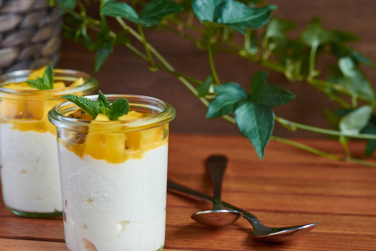 Zwei Gläser mit Solero-Dessert, gefüllt mit Joghurt, Mangostücken und Minzblättern, auf einem Holztisch.
