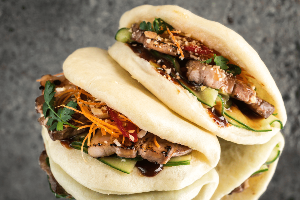 Drei gefüllte Bao Buns mit zartem Fleisch, Gurken, Karotten und Kräutern, angerichtet auf einem grauen Hintergrund.