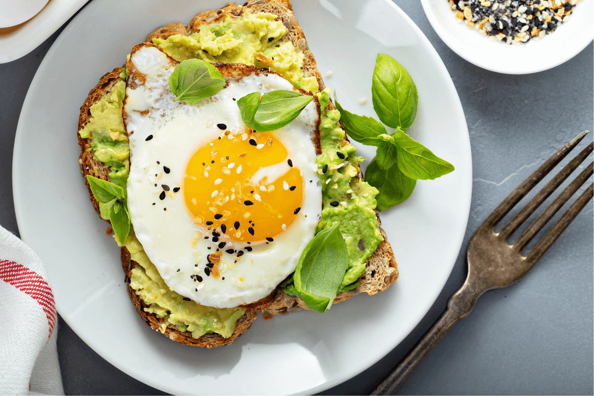 Avocado-Toast mit Spiegelei, garniert mit frischem Basilikum und schwarzen Sesamsamen auf einem weißen Teller.