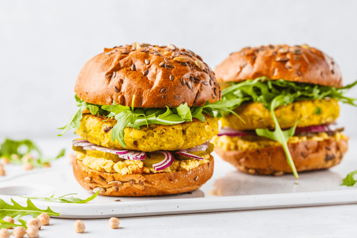 Zwei Kichererbsen-Walnuss-Burger auf einem weißen Teller, belegt mit Rucola und roten Zwiebeln in einem Brötchen mit Leinsamen.