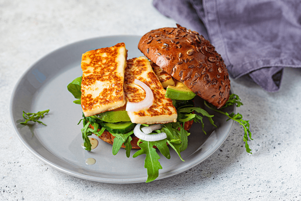 Halloumi-Burger auf einem grauen Teller, belegt mit frischem Rucola, Avocado, Zwiebelringen und einer Sriracha-Mayo.