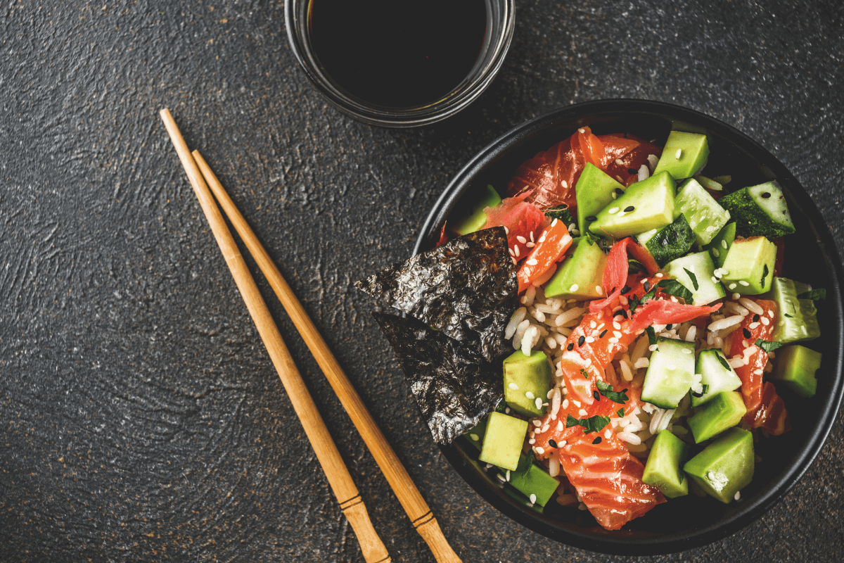 Schüssel mit Sushi Bowl, gefüllt mit Reis, Lachs, Gurken, Seetang und Sesam, neben Essstäbchen und Sojasauce.