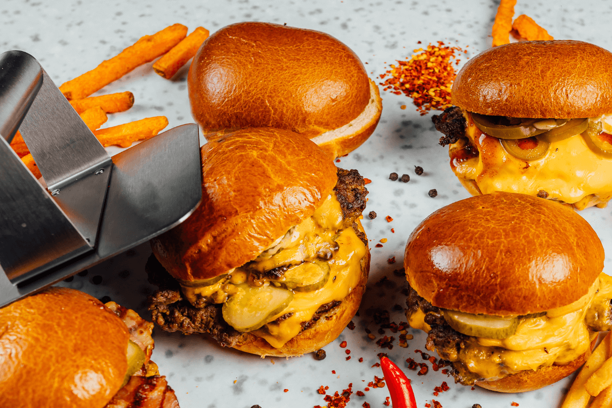 Smashed Burger auf einem Teller, umgeben von Süßkartoffel-Pommes, Gewürzen und einem Burger-Wender.