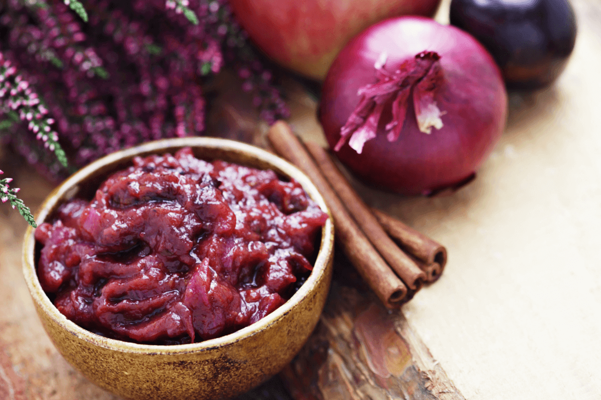 Schale mit Chutney aus Äpfeln und Zwiebeln, umgeben von Äpfeln, roten Zwiebeln, Zimtstangen und lila Früchten.