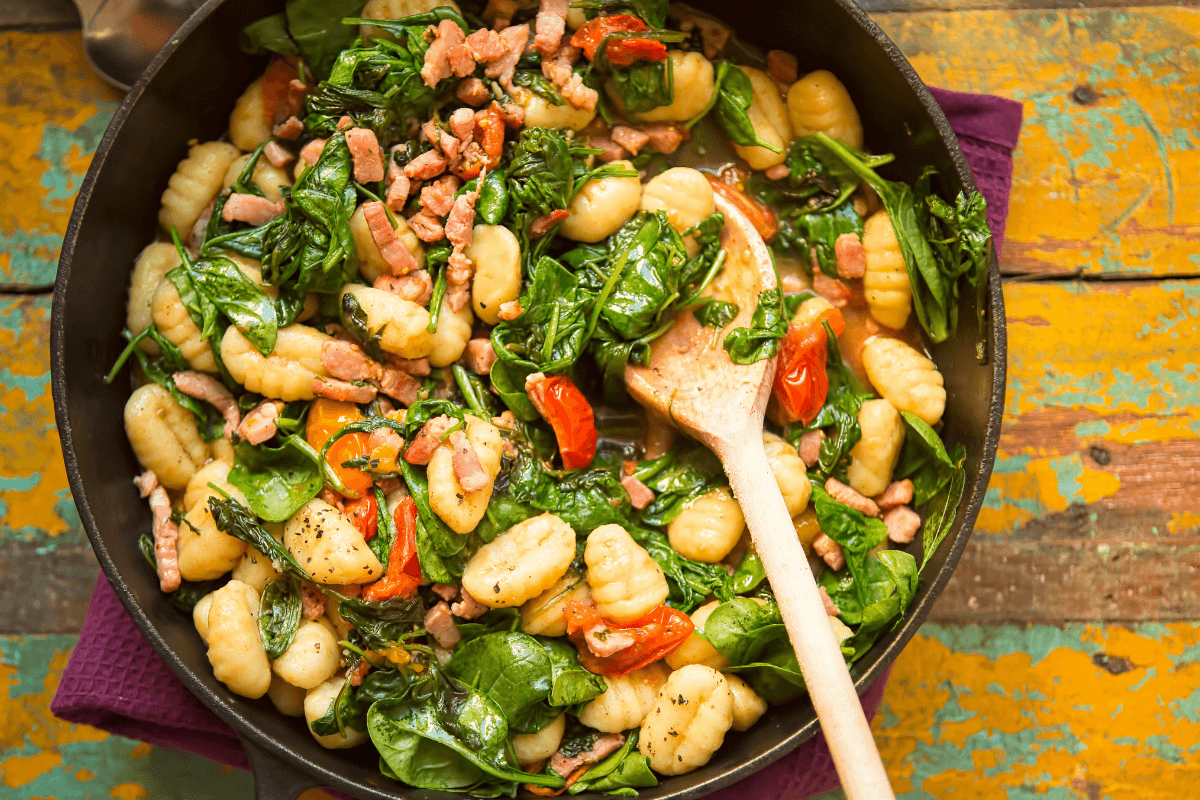 Bunte Tomaten-Gnocchi-Pfanne mit Spinat und Schinken, serviert in einer schwarzen Pfanne mit Holzlöffel.