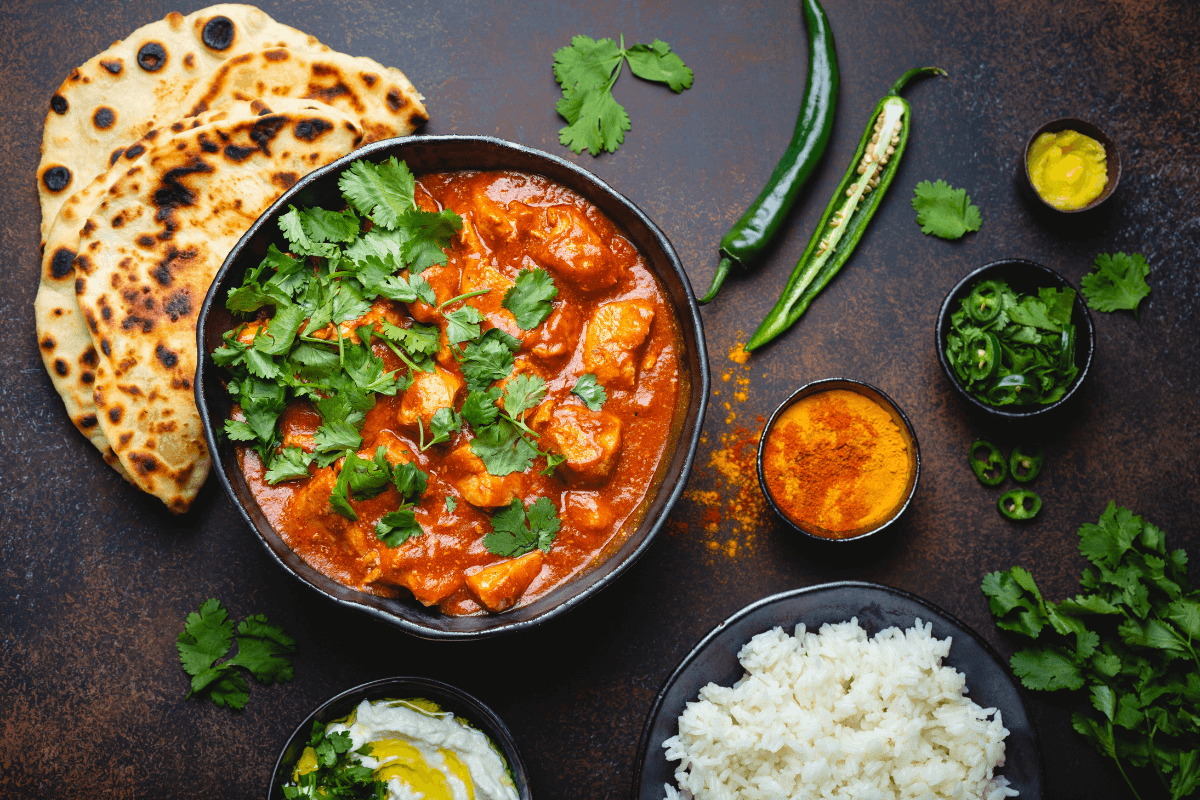 Schüssel mit Butter Chicken, garniert mit Koriander, dazu Naan-Brot, Reis, grüne Chilis und Gewürze auf einer dunklen Oberfläche.