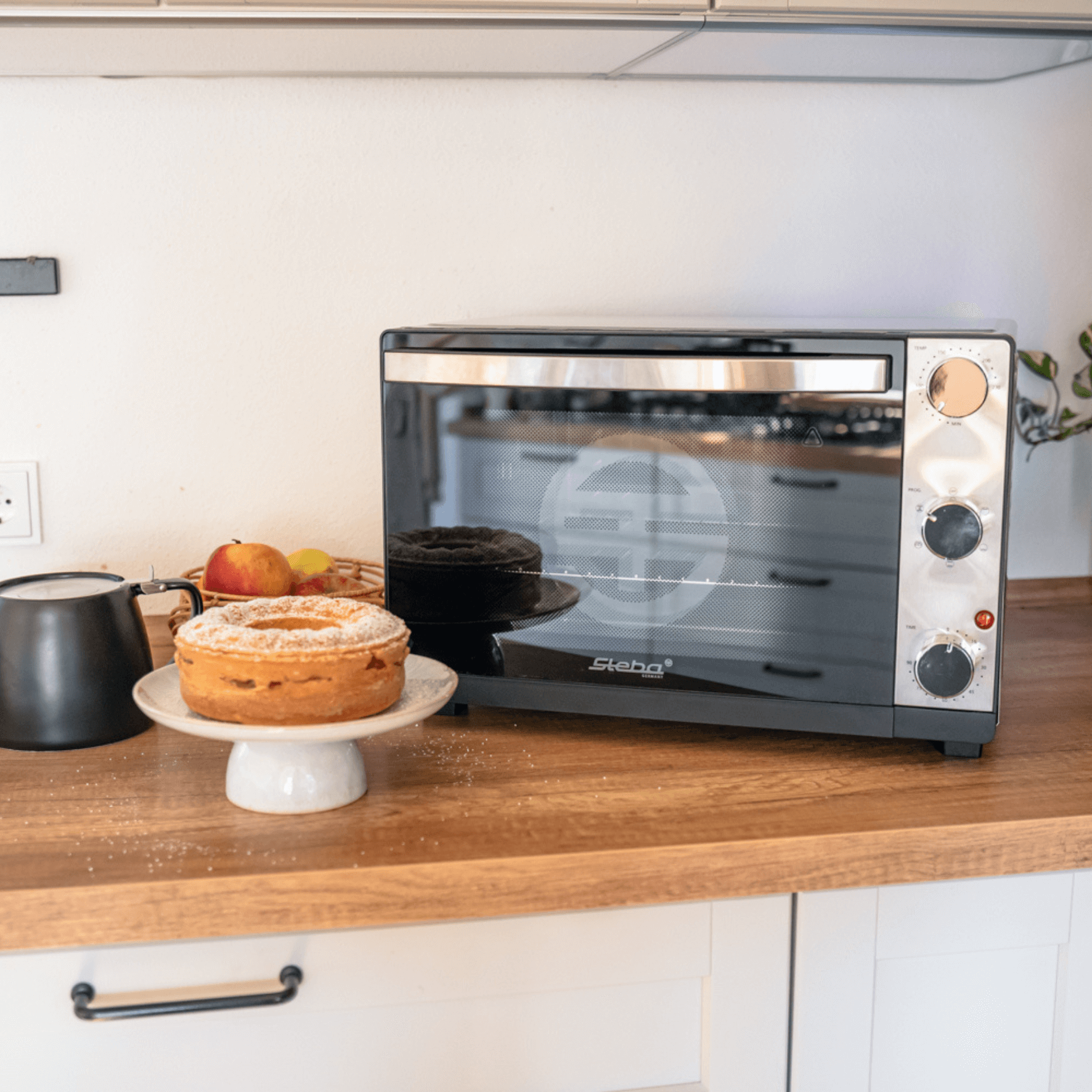 Grillbackofen Steba KB324 mit Edelstahl-Front, drei Drehreglern und Kuchen auf einem Teller auf einer Holzplatte.