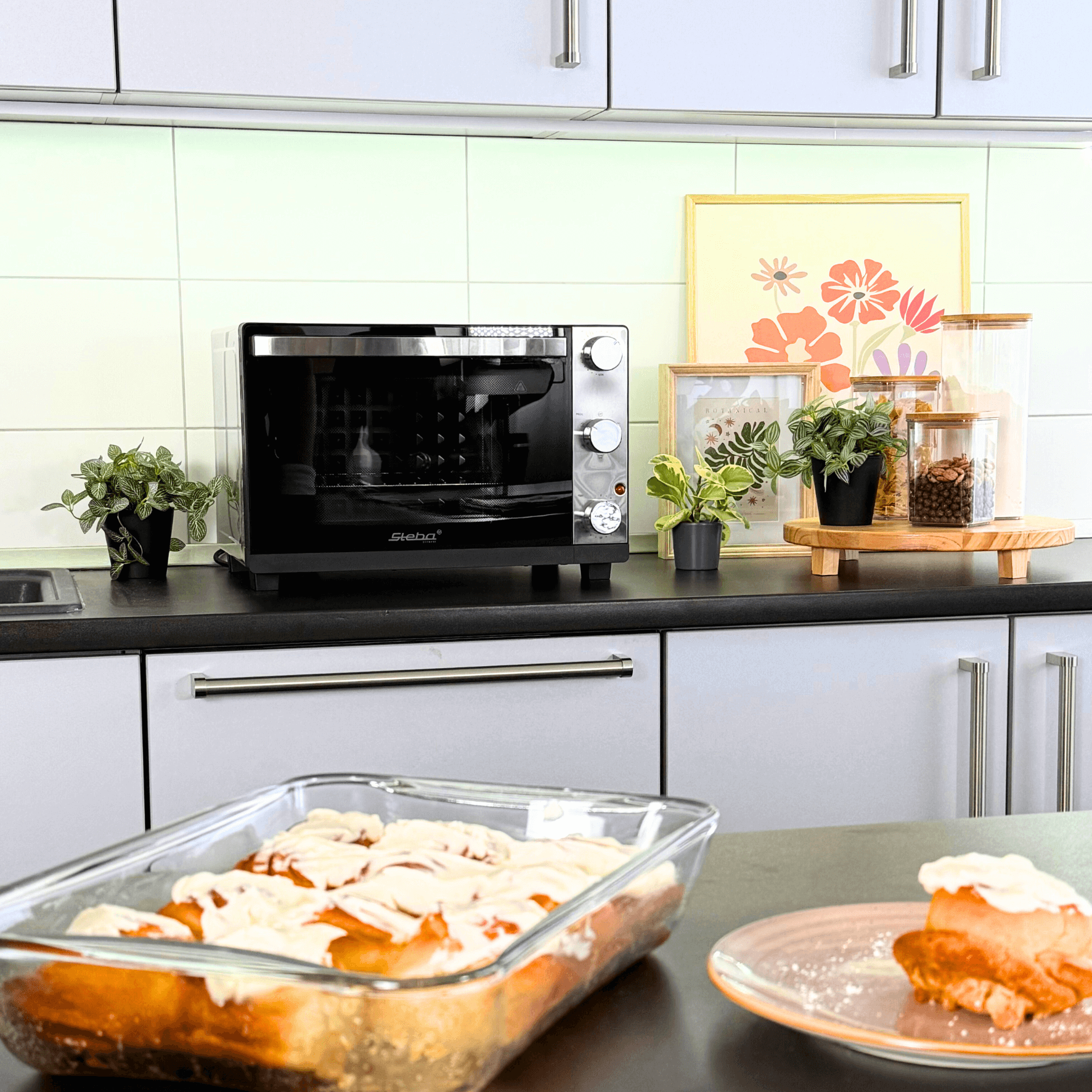 Grillbackofen Steba in Schwarz auf einer Küchenarbeitsplatte, umgeben von Pflanzen und einer Glasform mit Gebäck.
