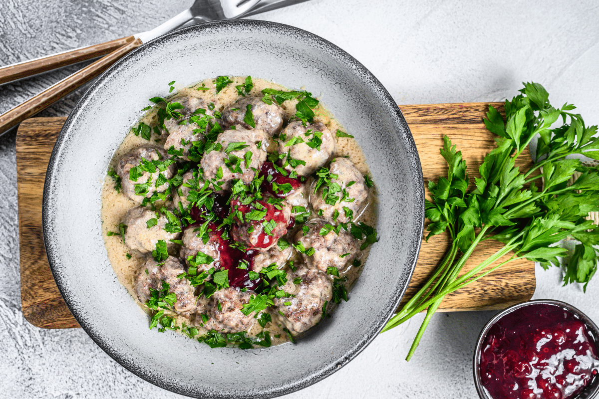 Köttbullar in einer cremigen Sauce, garniert mit frischer Petersilie und Preiselbeermarmelade, serviert in einer Schüssel.