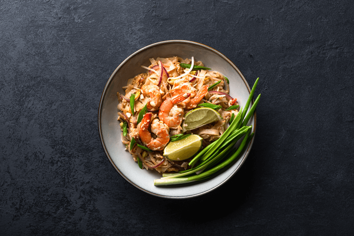 Schüssel mit Pad Thai, garniert mit Garnelen, Limettenscheiben, Erdnüssen und Frühlingszwiebeln auf dunklem Hintergrund.