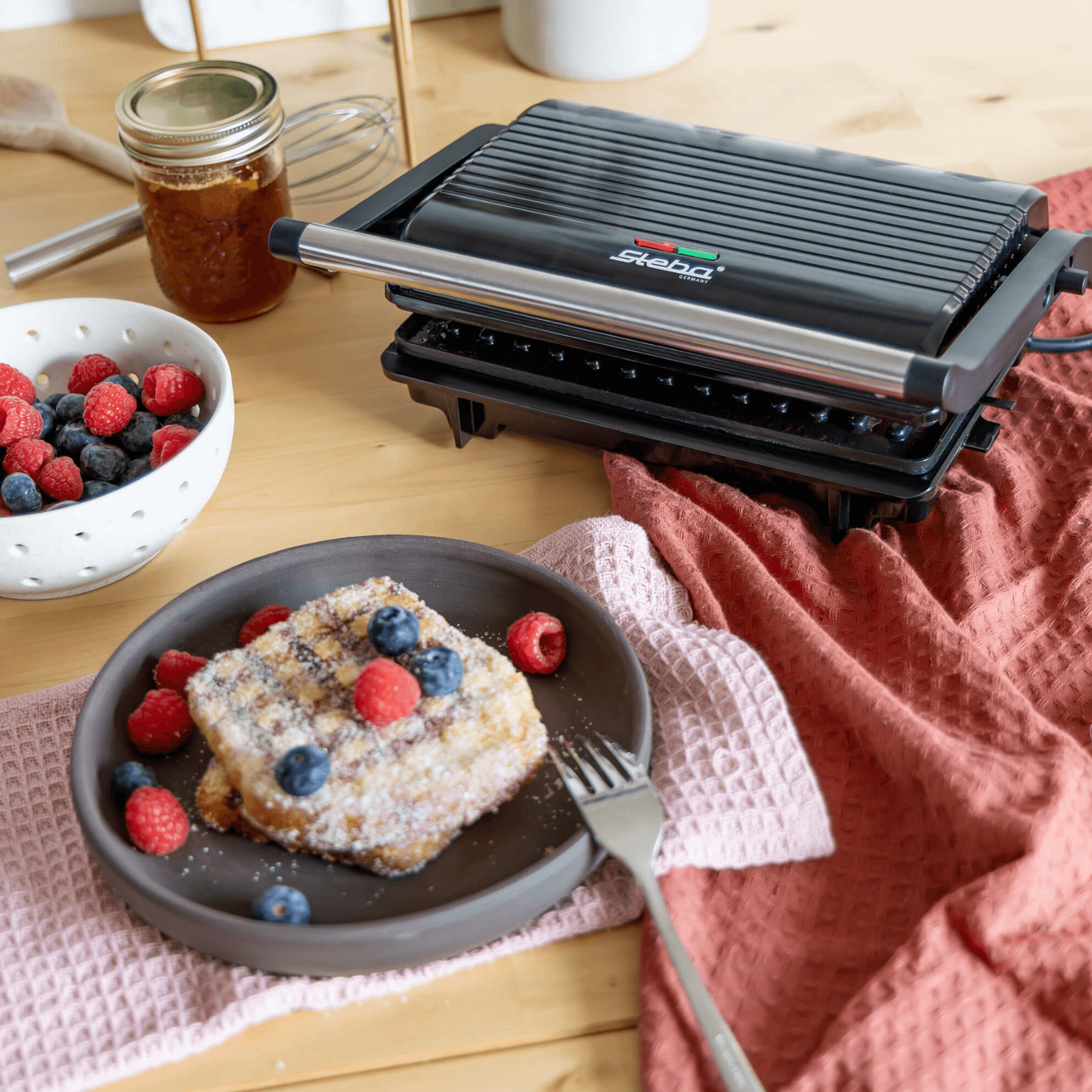 Kontaktgrill von Steba mit gerillter Oberfläche, daneben ein Teller mit süßem Toast und frischen Beeren sowie ein Glas Marmelade.