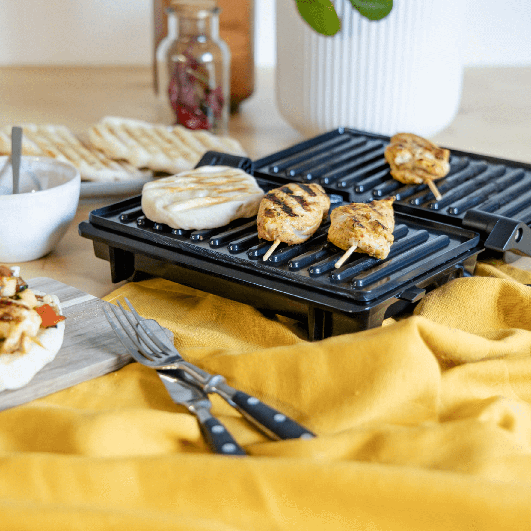 Kontaktgrill von Steba mit Antihaftbeschichtung, gegrilltem Hähnchen und Fladenbrot, auf einem Tisch mit gelber Decke.