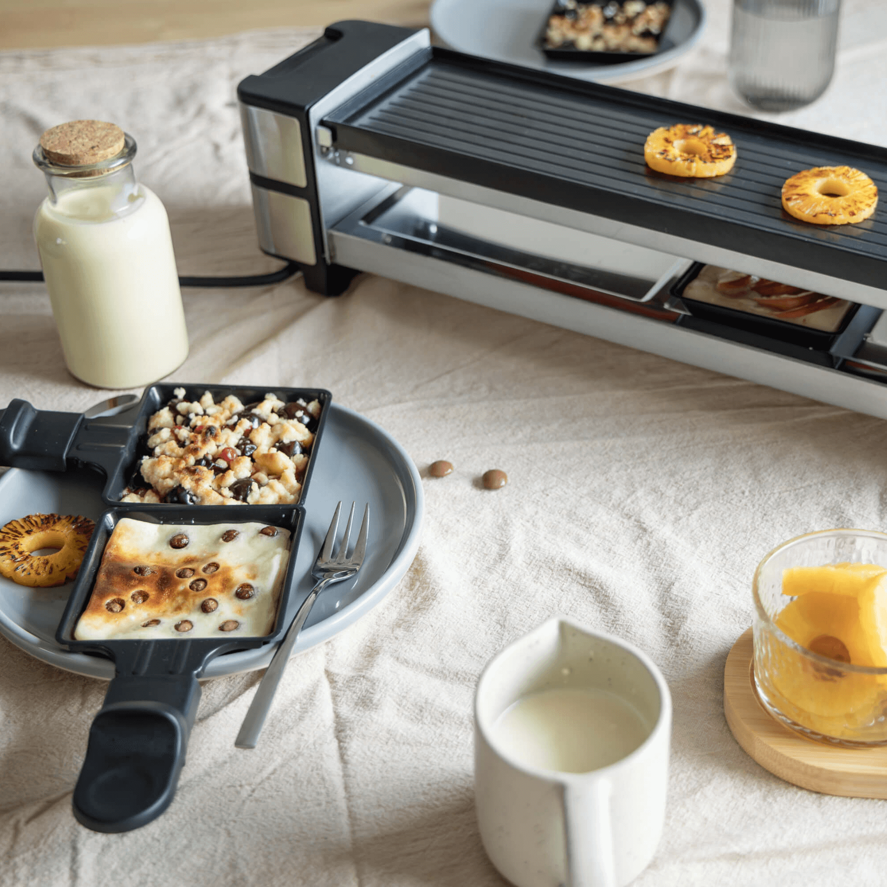 Raclette-Grill von Steba mit zwei Pfännchen, gegrilltem Obst und einer Flasche Milch auf einem Tisch.